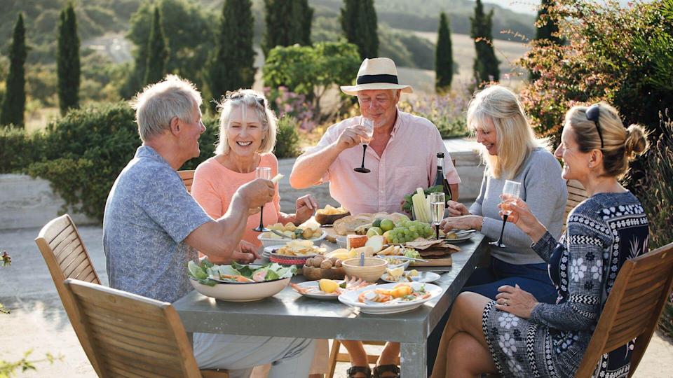 A group of mature friends are sitting around an outdoor dining table, eating and drinking.