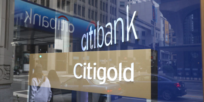 Two blue signs and a golden sign, all with the Citi logo, are seen reflected in front of a bank branch in San Francisco.